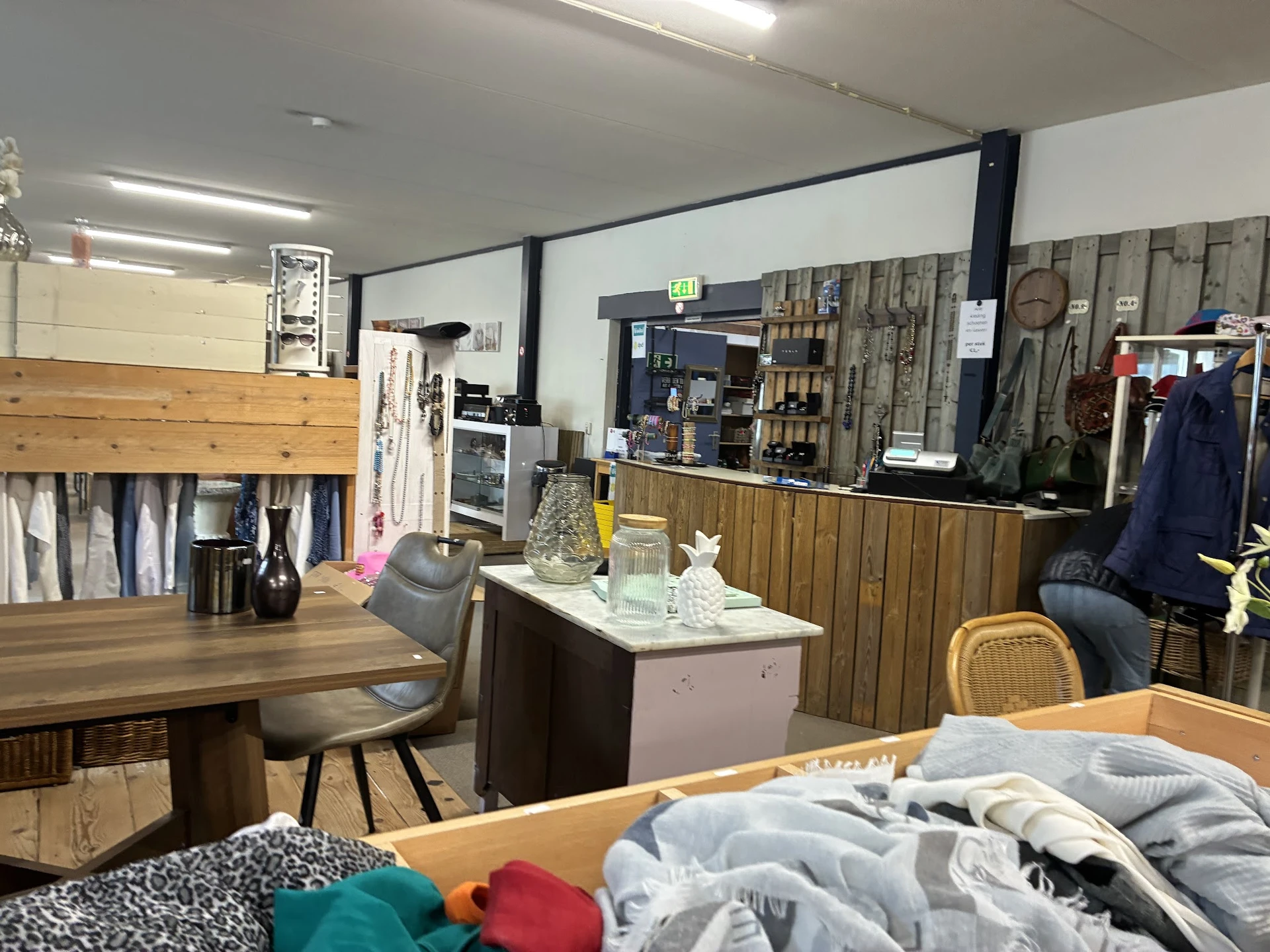 Binnenkant van kringloopwinkel met houten tafels, kleding op rekken, stellingen met spullen en diverse tweedehands artikelen