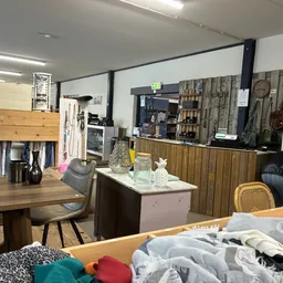 Binnenkant van kringloopwinkel met houten tafels, kleding op rekken, stellingen met spullen en diverse tweedehands artikelen