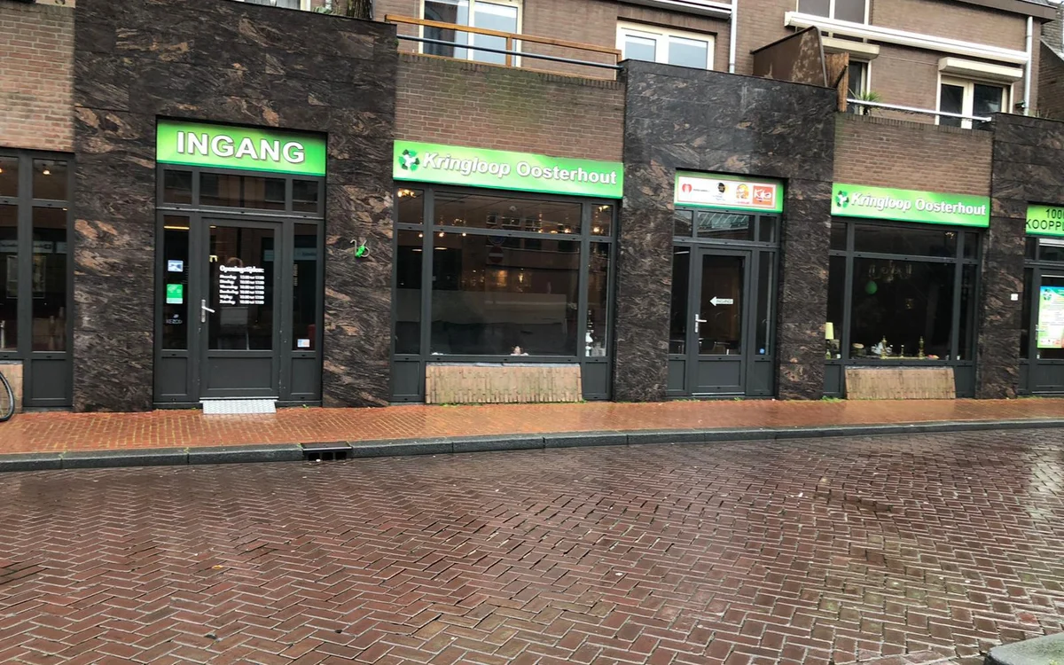 Voorgevel van kringloopwinkel met donkere stenen pui, groene INGANG-borden en grote etalageramen aan een plein met straatstee