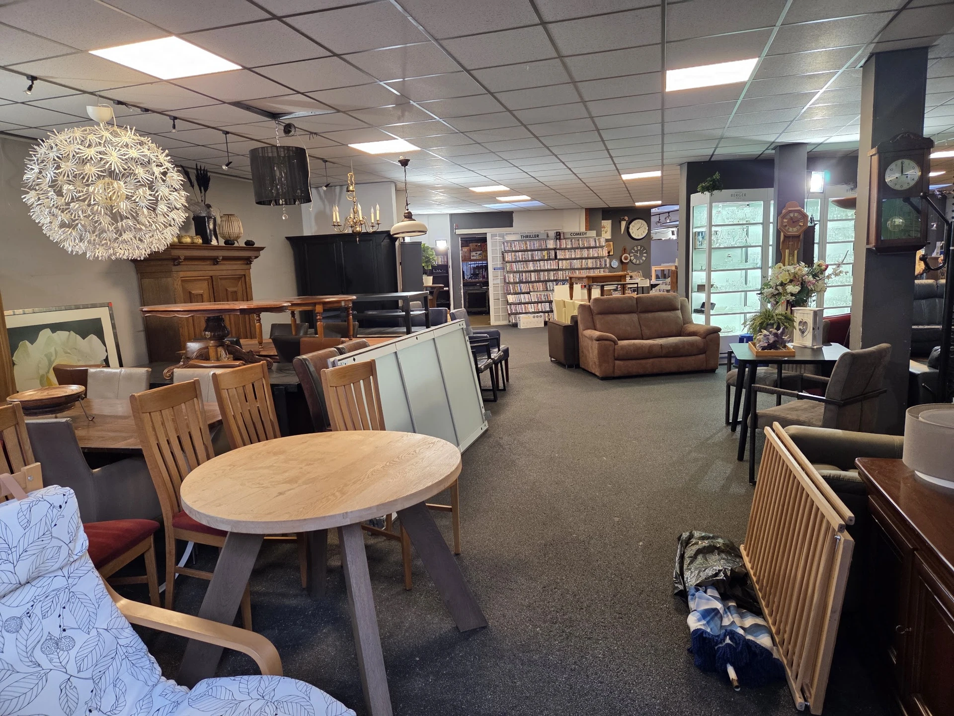 Ruime winkelruimte met ronde houten eettafel met stoelen, bruine fauteuil, televisie, stellingen met spullen en varend licht