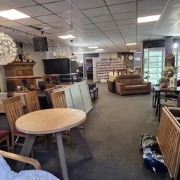 Ruime winkelruimte met ronde houten eettafel met stoelen, bruine fauteuil, televisie, stellingen met spullen en varend licht