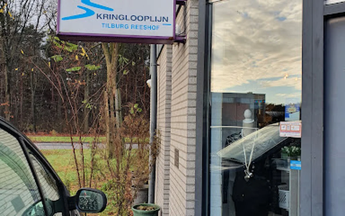 Gevel van Kringlooplijn Tilburg Reeshof met bord en glazen deur, parkeerplaats zichtbaar