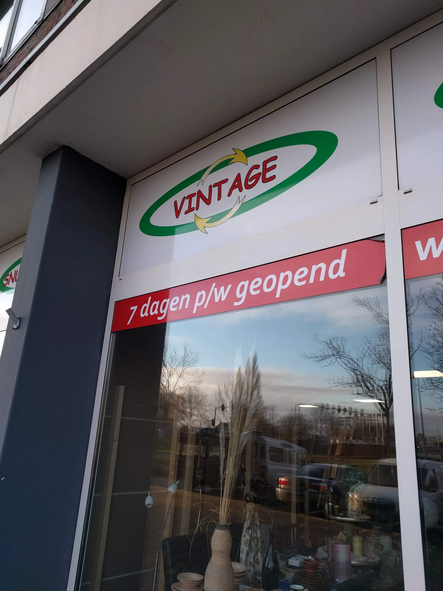 Winkelruit met VINTAGE logo en rode banner '7 dagen p/w geopend', winkelstraat zichtbaar in reflectie