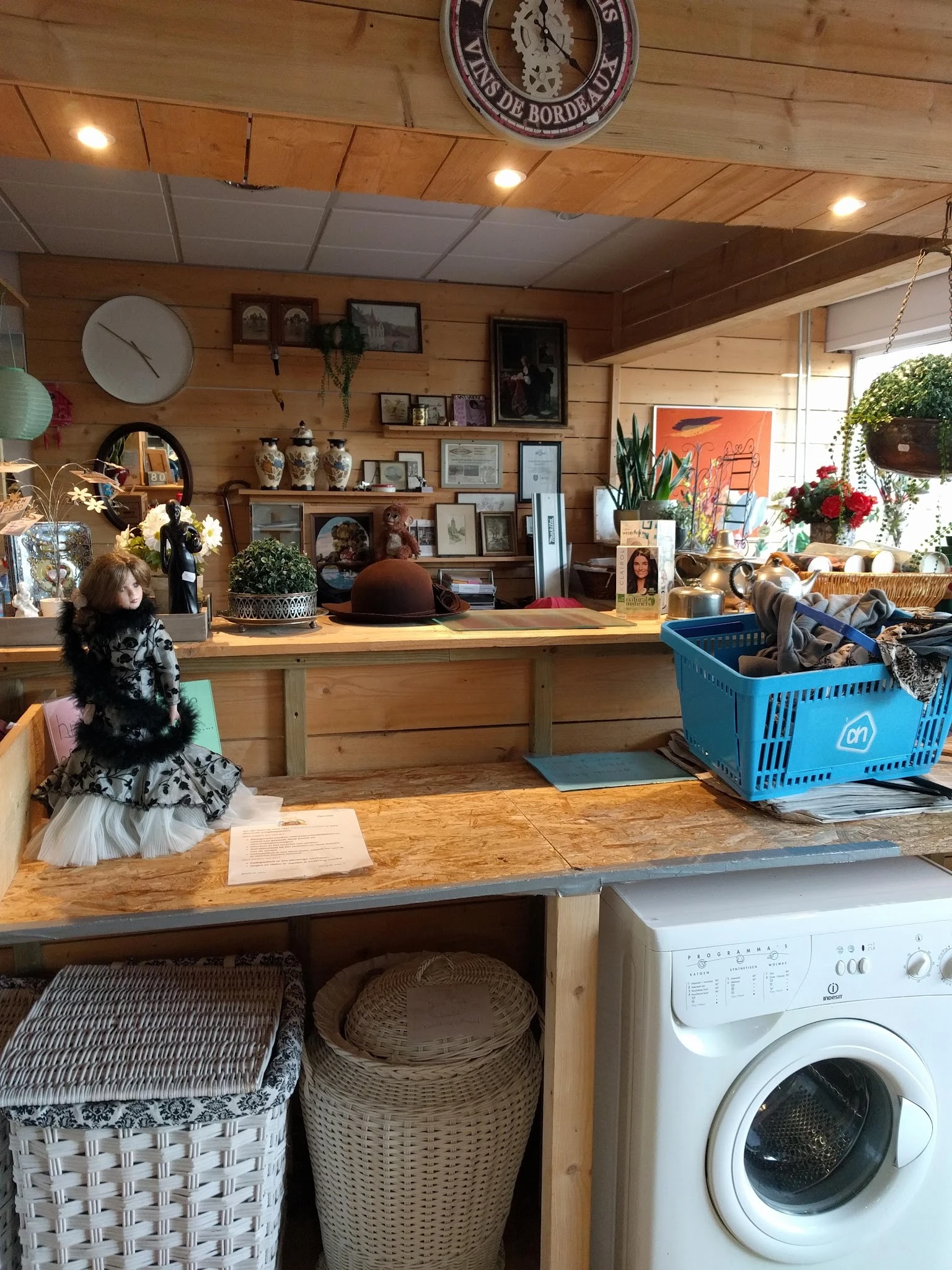 Houten toonbank van kringloopwinkel met daarop diverse spullen, achter de balie stellingen met decoratie, bloemen en huisraad