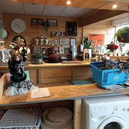 Houten toonbank van kringloopwinkel met daarop diverse spullen, achter de balie stellingen met decoratie, bloemen en huisraad