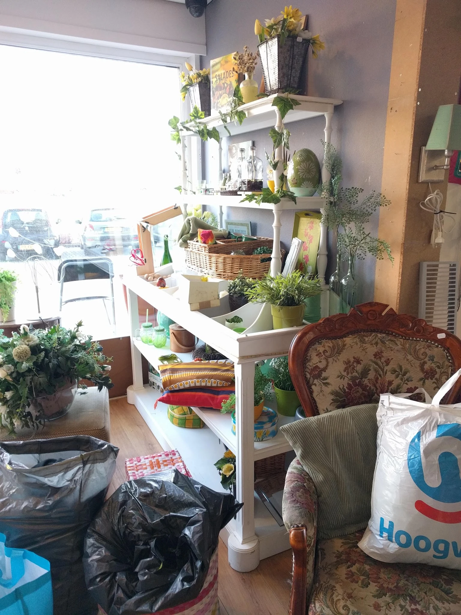 Winkelinterieur met witte stellingen vol plantenpotten, manden, boeken en huishoudartikelen; rode fauteuil op de voorgrond