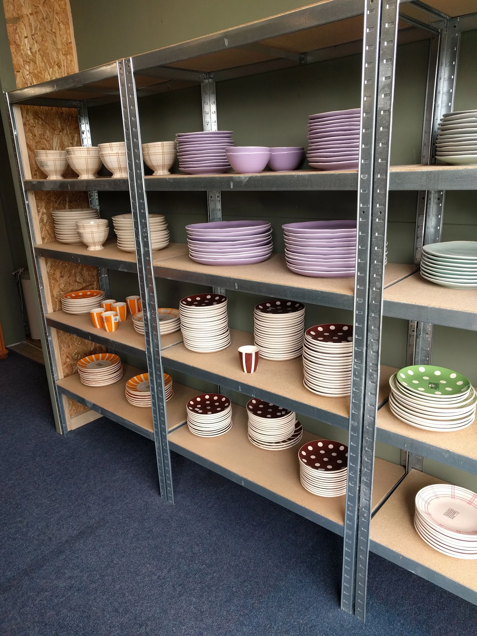 Stellingen met uitgestalde serviesgoed: stapels borden en schalen in verschillende kleuren waaronder paars, wit, bruin en gro