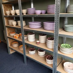Stellingen met uitgestalde serviesgoed: stapels borden en schalen in verschillende kleuren waaronder paars, wit, bruin en gro