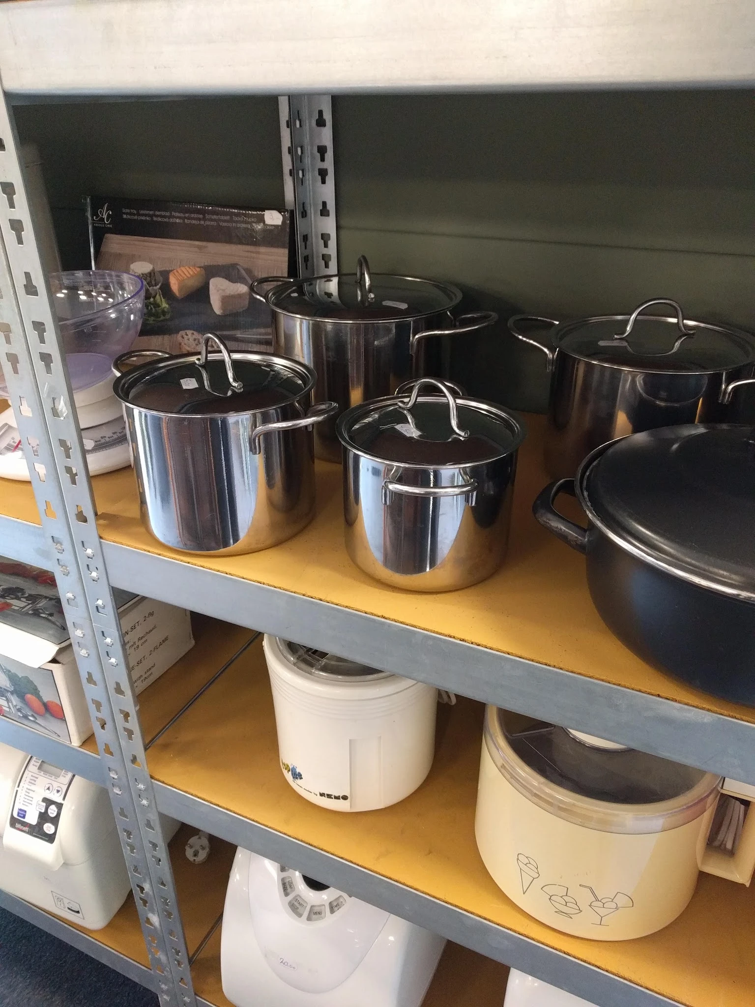 Verschillende roestvrijstalen pannen en potten met deksels op een geel schap in de kringloopwinkel