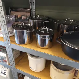 Verschillende roestvrijstalen pannen en potten met deksels op een geel schap in de kringloopwinkel