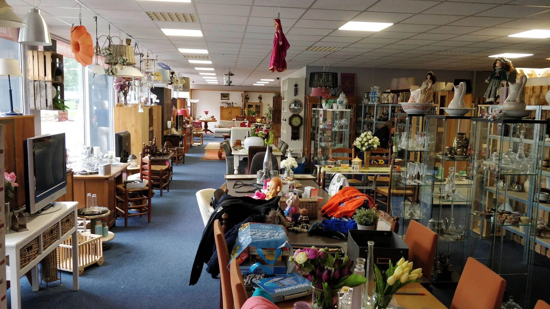 Brede blik op de winkelvloer met diverse tweedehands spullen, meubels, decoraties en stellingen langs de zijkanten.