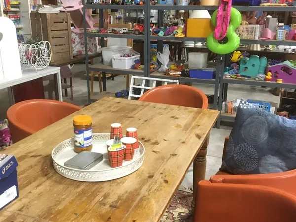 Houten tafel met oranje stoelen in een kringloopwinkel vol stellingen met spullen op de achtergrond