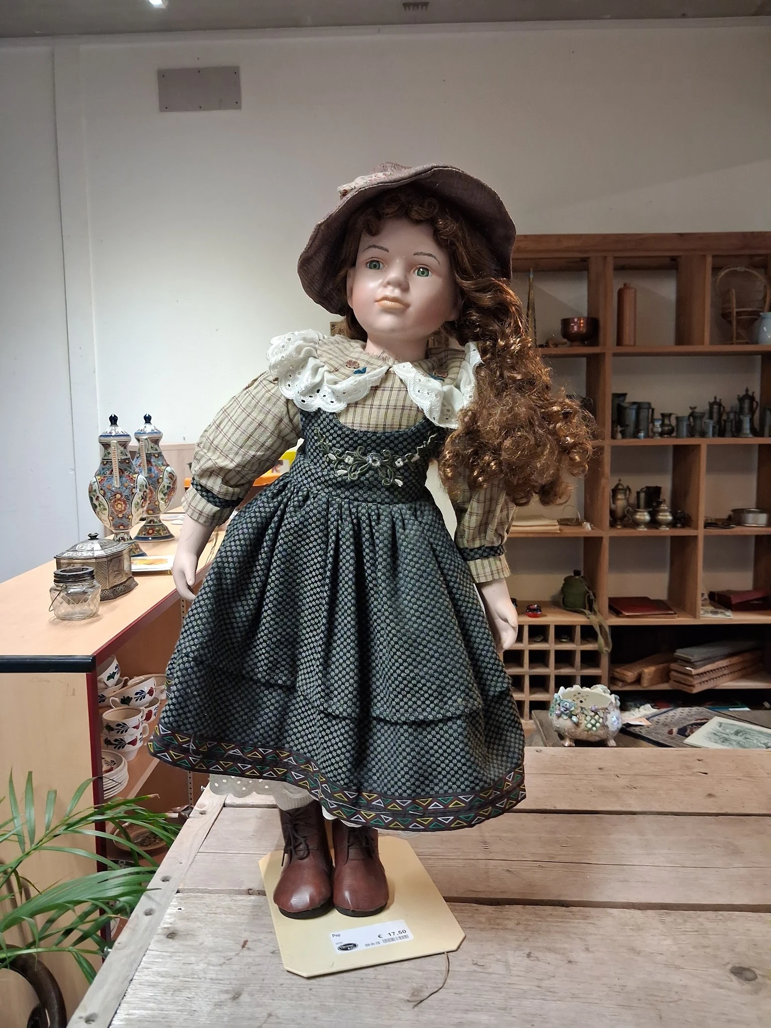 Porseleinenpop in historische jurk met kant en geruit patroon, staand op tafel in een winkelinterieur