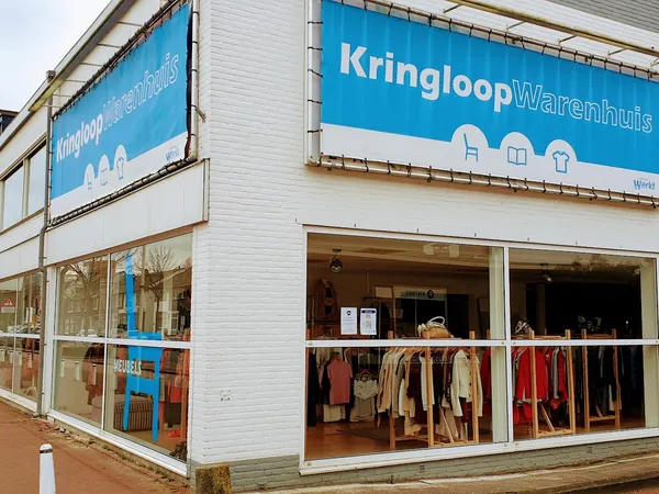 Witte gevel van Kringloopwarenhuis met blauwe uithangborden, grote etalagevensters met tweedehands kleding zichtbaar