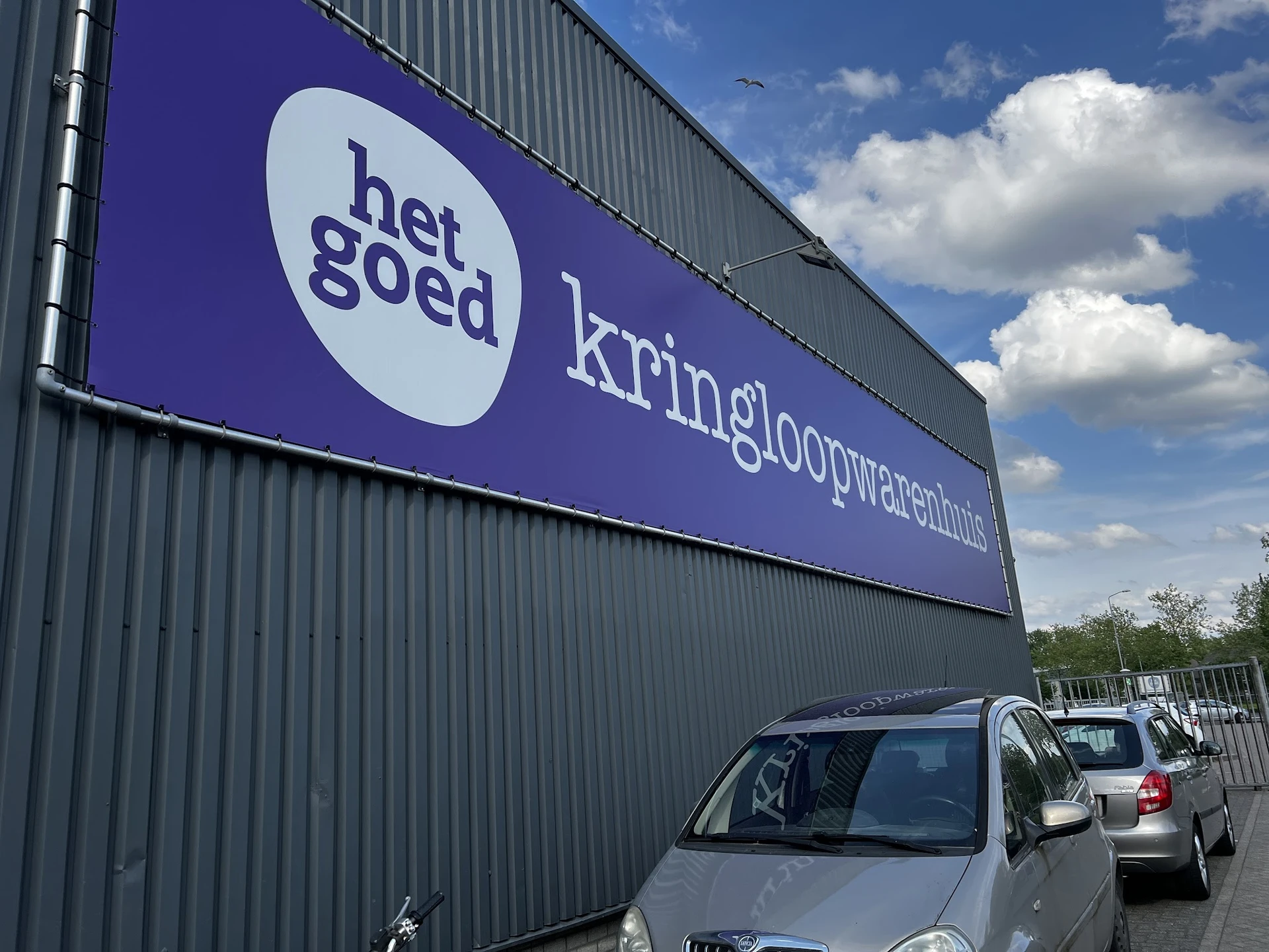 Gevel van kringloopwinkel met paars bord met 'het goed' logo en 'kringloopwarenhuis' tekst, geparkeerde auto's ervoor