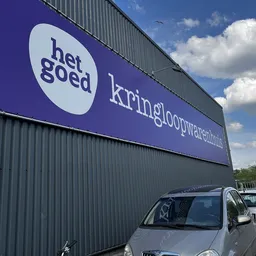 Gevel van kringloopwinkel met paars bord met 'het goed' logo en 'kringloopwarenhuis' tekst, geparkeerde auto's ervoor