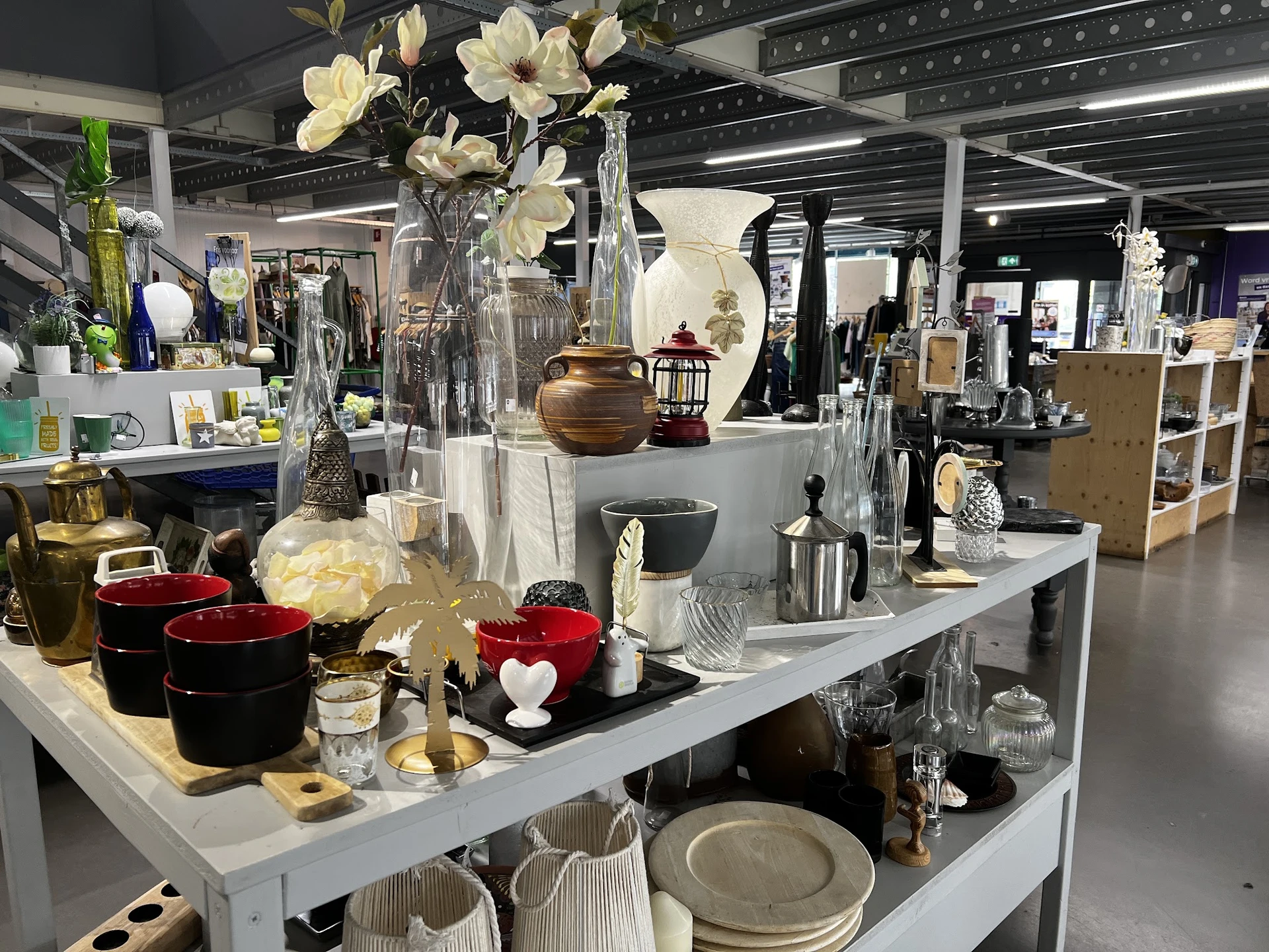 Uitgestalde huishoudelijke artikelen en decoratie op witte tafels, waaronder vazen met bloemen, rode kaarsen, servies en acce
