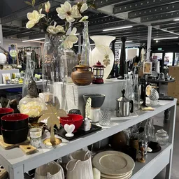 Uitgestalde huishoudelijke artikelen en decoratie op witte tafels, waaronder vazen met bloemen, rode kaarsen, servies en acce