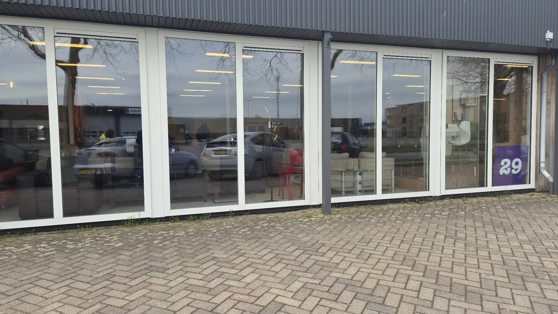 Glazen winkelpui met witte frames en grote ramen, parkeerplaats met auto's zichtbaar in reflectie