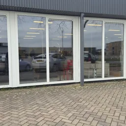 Glazen winkelpui met witte frames en grote ramen, parkeerplaats met auto's zichtbaar in reflectie