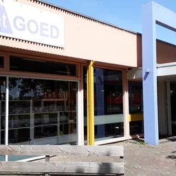 Gevel van kringloopwinkel met witte en bruine gevelbekleding, grote ramen en gele steunpilaar, winkelname 'Het GOED' gedeelte
