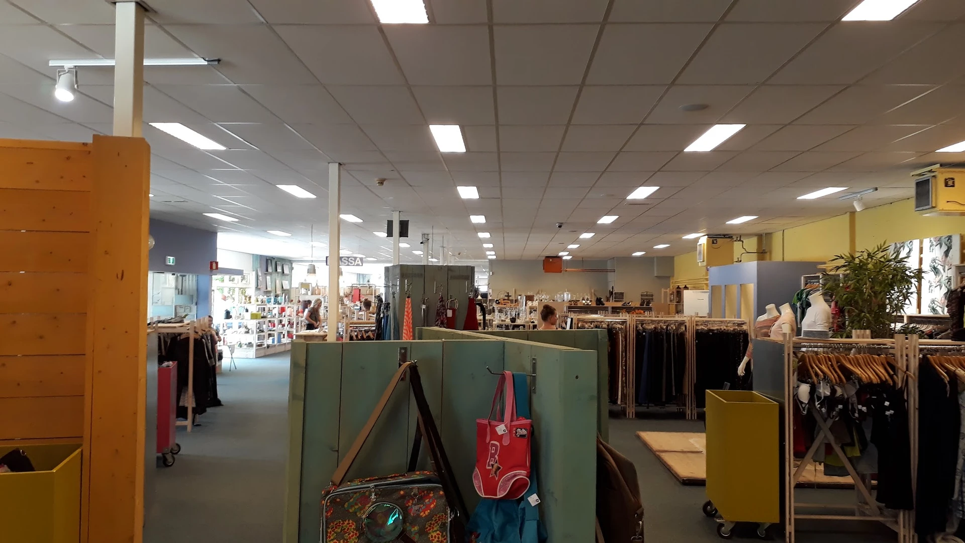 Overzicht van de winkelvloer met kledingrekken, tassen en diverse tweedehands artikelen onder fluorescente verlichting