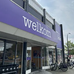 Paarse gevel van kringloopwinkel met grote ramen en winkelnaam 'Welkom' in witte letters