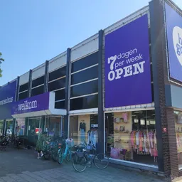 Gevel van kringloopwinkel met paarse borden, 'welkom' tekst, grote ramen met uitstallingen en fietsen geparkeerd ervoor