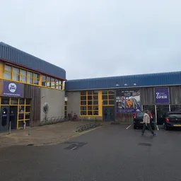 Voorgevel van kringloopwarenhuis met gele deuren en ramen, parkeerplaats met enkele auto's en personen voor de ingang