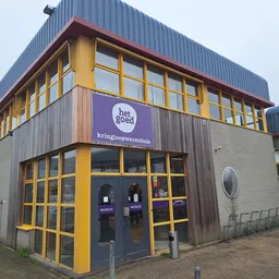 Gevel van kringloopwinkel met geel-zwart frame, grote ramen en paars logo 'het goed'