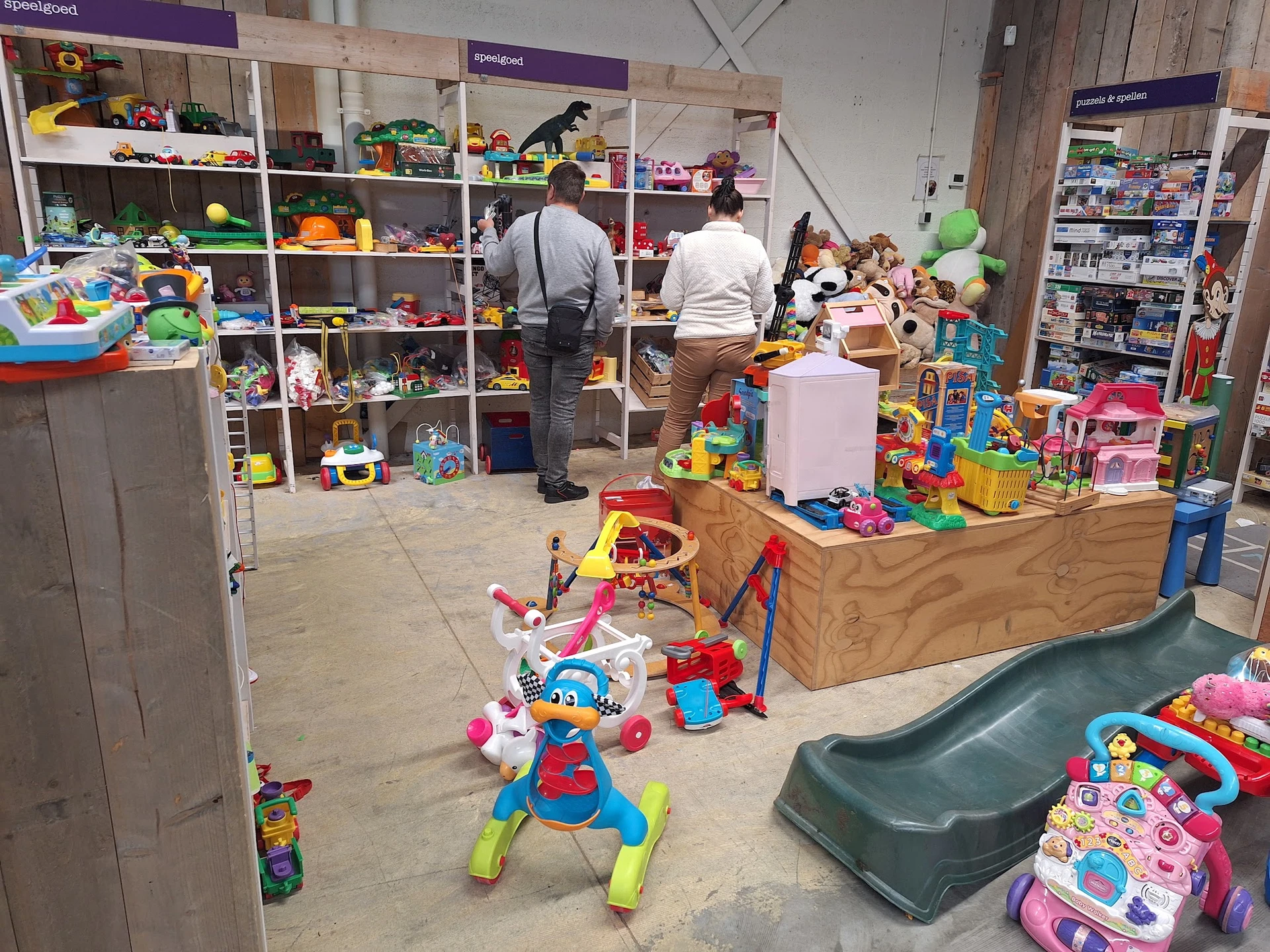 Speelgoedafdeling in kringloopwinkel met speelgoed uitgestald op houten display en stellingen met twee medewerkers in het mid