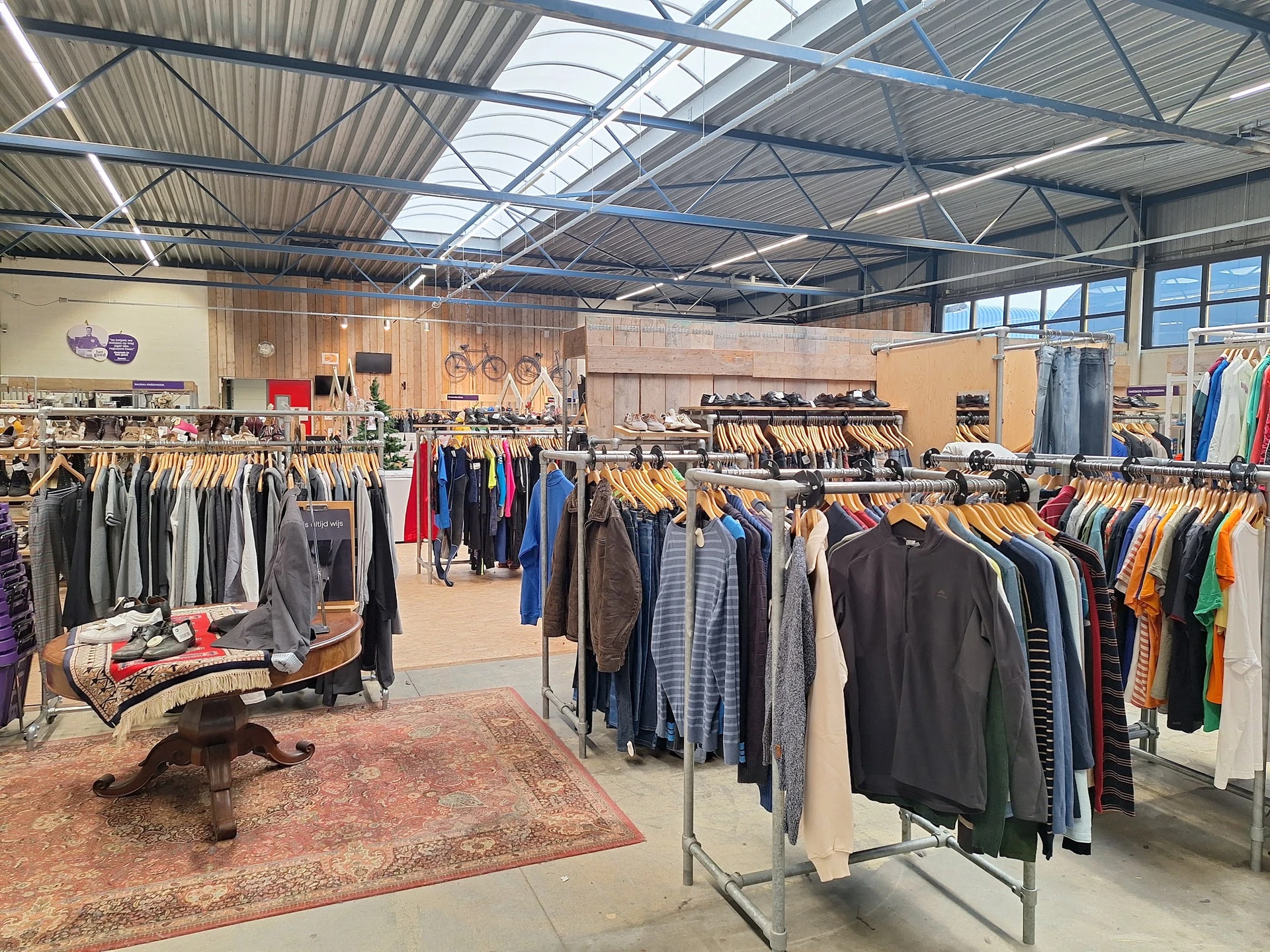 Ruim winkelinterieur met kledingrekken vol tweedehands kleding, een verzameling tafels en een gezellige winkelsfeer.