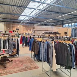 Ruim winkelinterieur met kledingrekken vol tweedehands kleding, een verzameling tafels en een gezellige winkelsfeer.
