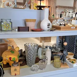 Uitgestalde huishoudarikelen en decoratie op twee witte planken, waaronder vazen, glazen, een gele spaarpot en verschillende 