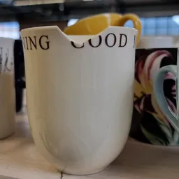 Witte vaas of kan met gele hengsels en gedeeltelijk zichtbare tekst, tussen andere serviesgoed op een plank