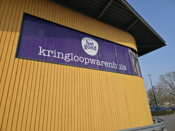 Gele gevel van kringloopwarenhuis met paars bord waarop winkelnaam in wit en logo