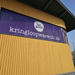 Gele gevel van kringloopwarenhuis met paars bord waarop winkelnaam in wit en logo