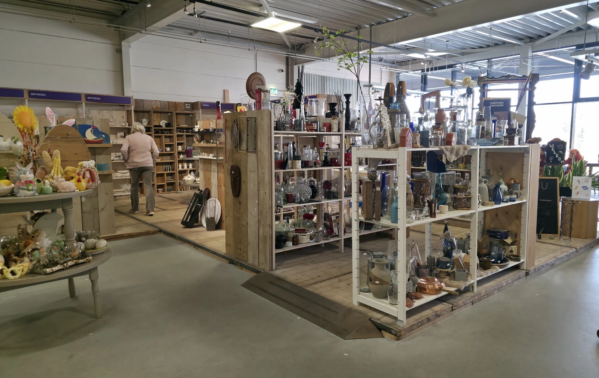 Overzicht van kringloopwinkel met houten en witte stellingen vol huishoudartikelen, decoratie en diverse goederen