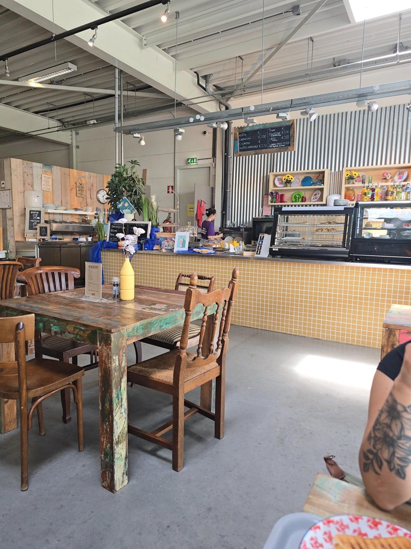 Houten tafels met stoelen in een lunchhoek, gele tegelwand achter de toonbank met koffieapparatuur en kasten met producten