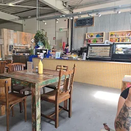 Houten tafels met stoelen in een lunchhoek, gele tegelwand achter de toonbank met koffieapparatuur en kasten met producten