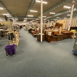 Spacious warehouse interior with wooden furniture, storage boxes, and various secondhand items displayed throughout the sales