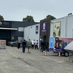 Exterieur van kringloopwinkel met bezoekers en promotiewagen op parkeerterrein