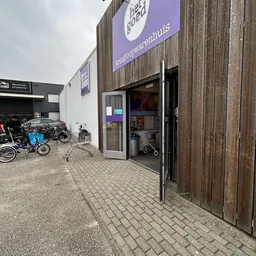 Ingang van kringloopwinkel met paarse uithangbord, houten deuren en stellingen met tweewielers buiten