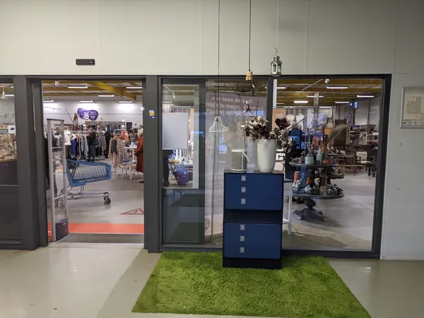 Entree van kringloopwinkel met glazen deuren, groene loopmat en blauwe display-kastjes, zicht op winkelruimte met artikelen e