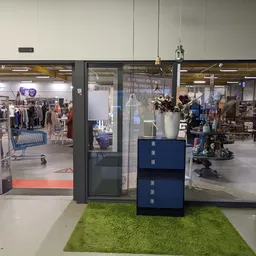 Entree van kringloopwinkel met glazen deuren, groene loopmat en blauwe display-kastjes, zicht op winkelruimte met artikelen e