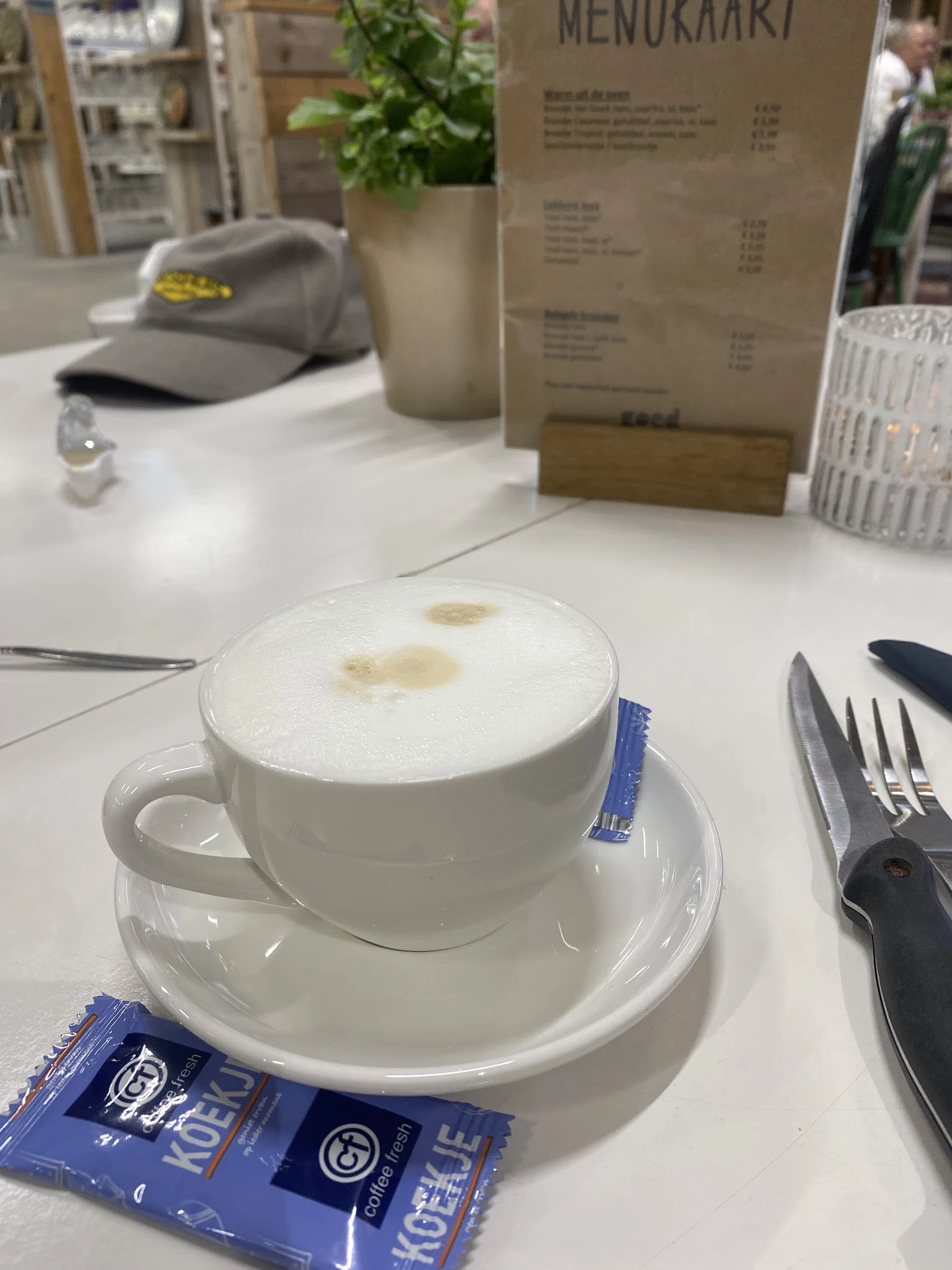 Kopje cappuccino met melkschuim op een witte schotel, met bestek en tafelgerei
