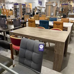 Houten eettafel met gekleurde stoelen in uitstalling, diverse meubels zichtbaar op achtergrond
