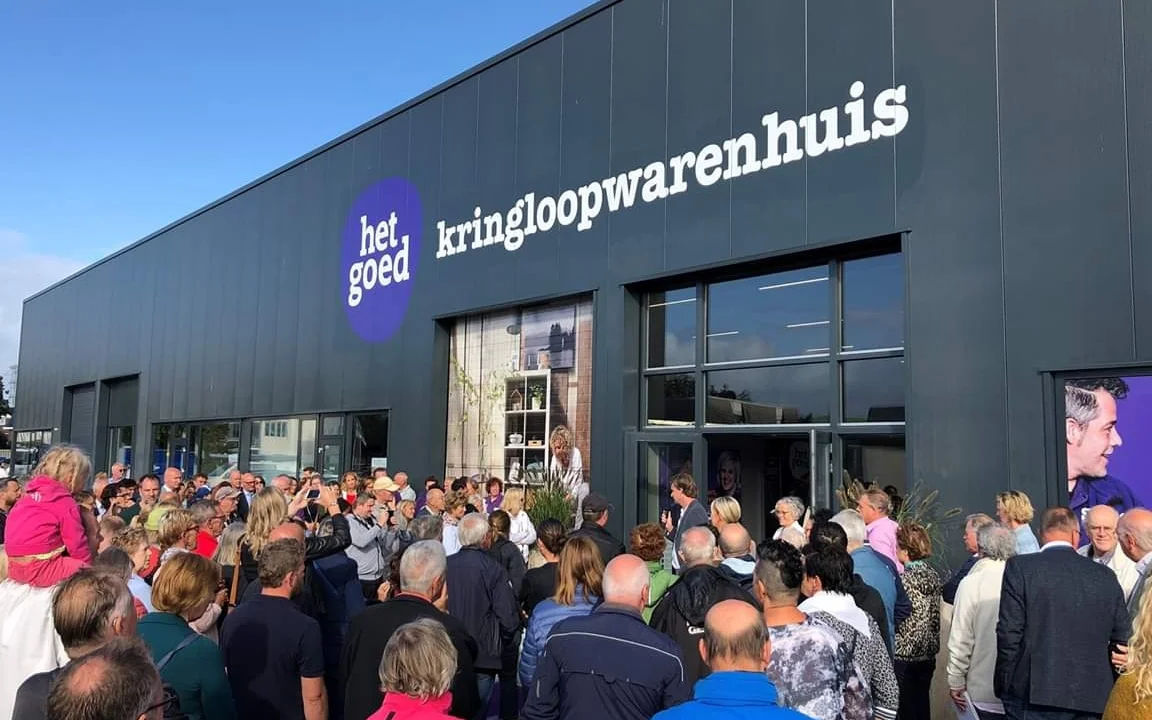 Gevel van kringloopwinkel met neonlogo en grote groep bezoekers bij de ingang