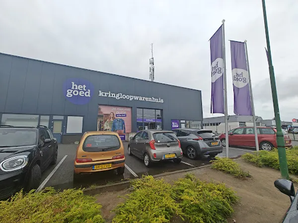 Grijze gevel van kringloopwinkel Het Goed met paarse banners en geparkeerde auto's op het parkeerterrein