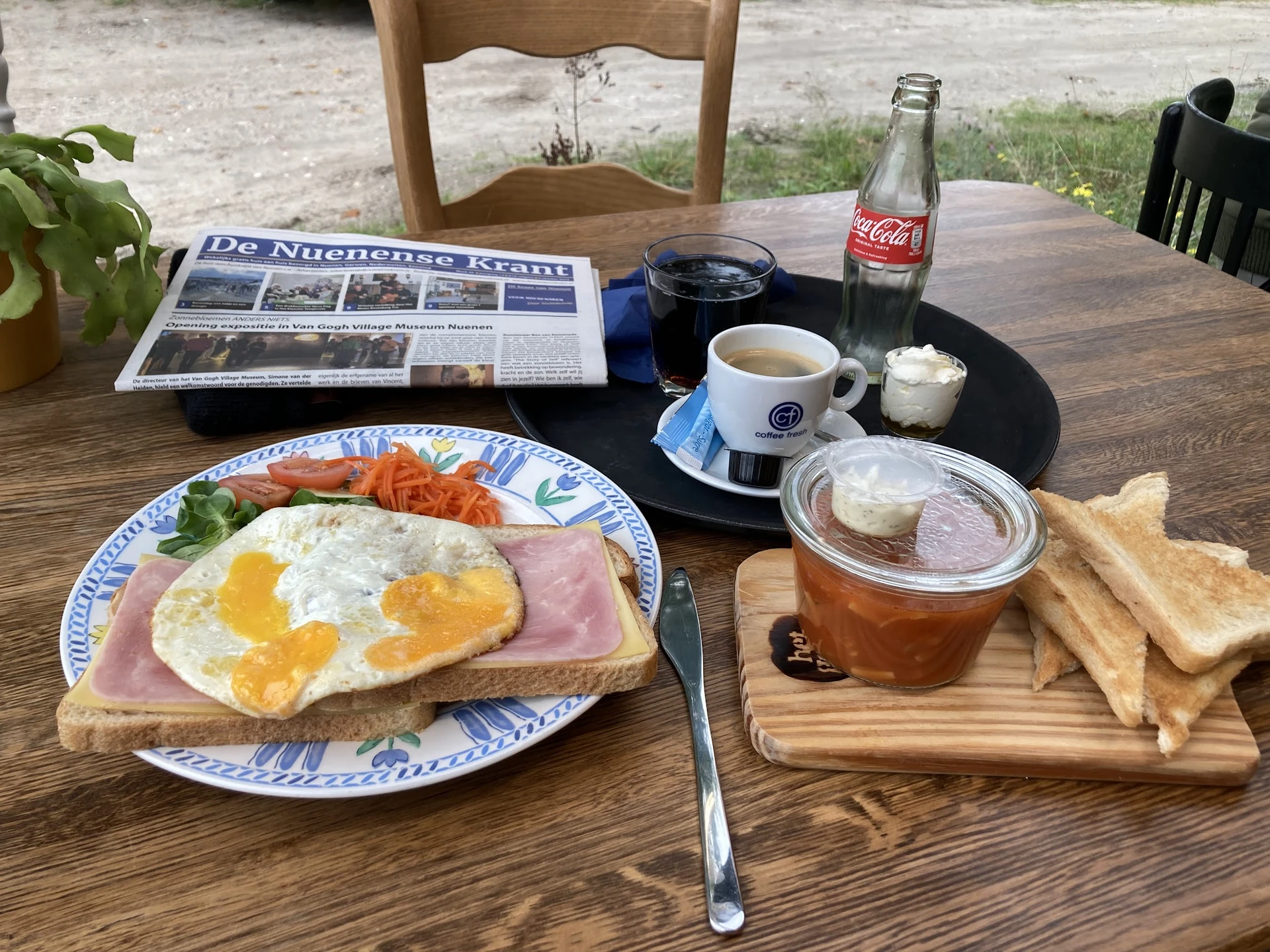 Ontbijtbord met spiegelei, ham en toast op een terras, met koffie en Coca-Cola